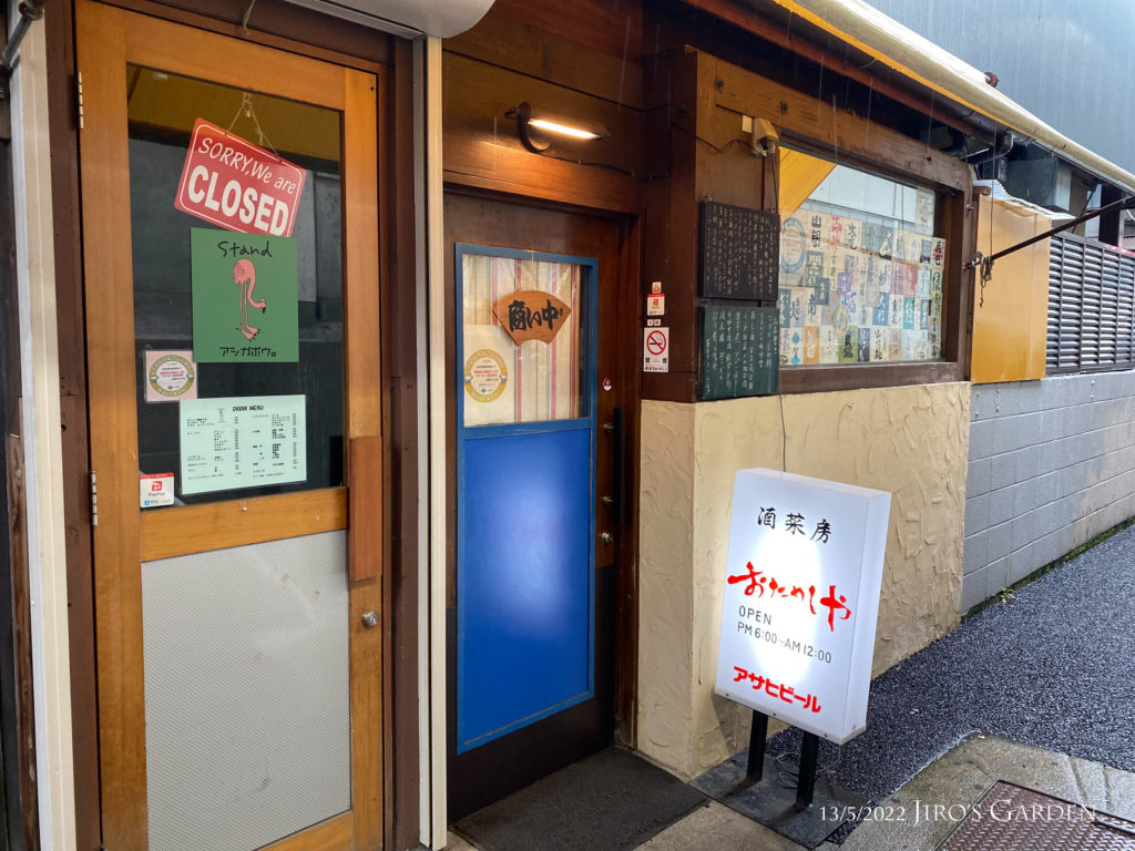 店舗正面を左斜め前から写す。青いドアとガラス一面に貼られた日本酒のラベル。白い電飾看板。