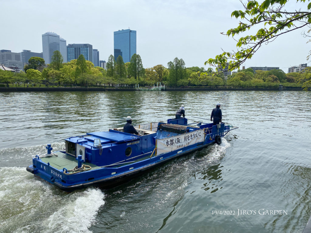 白い波をたてながら川をゆく清掃用の青い船舶。12×3mくらいか。3名の乗組員。「水都大阪」川をきれいに、のバナーが側面に貼られている。